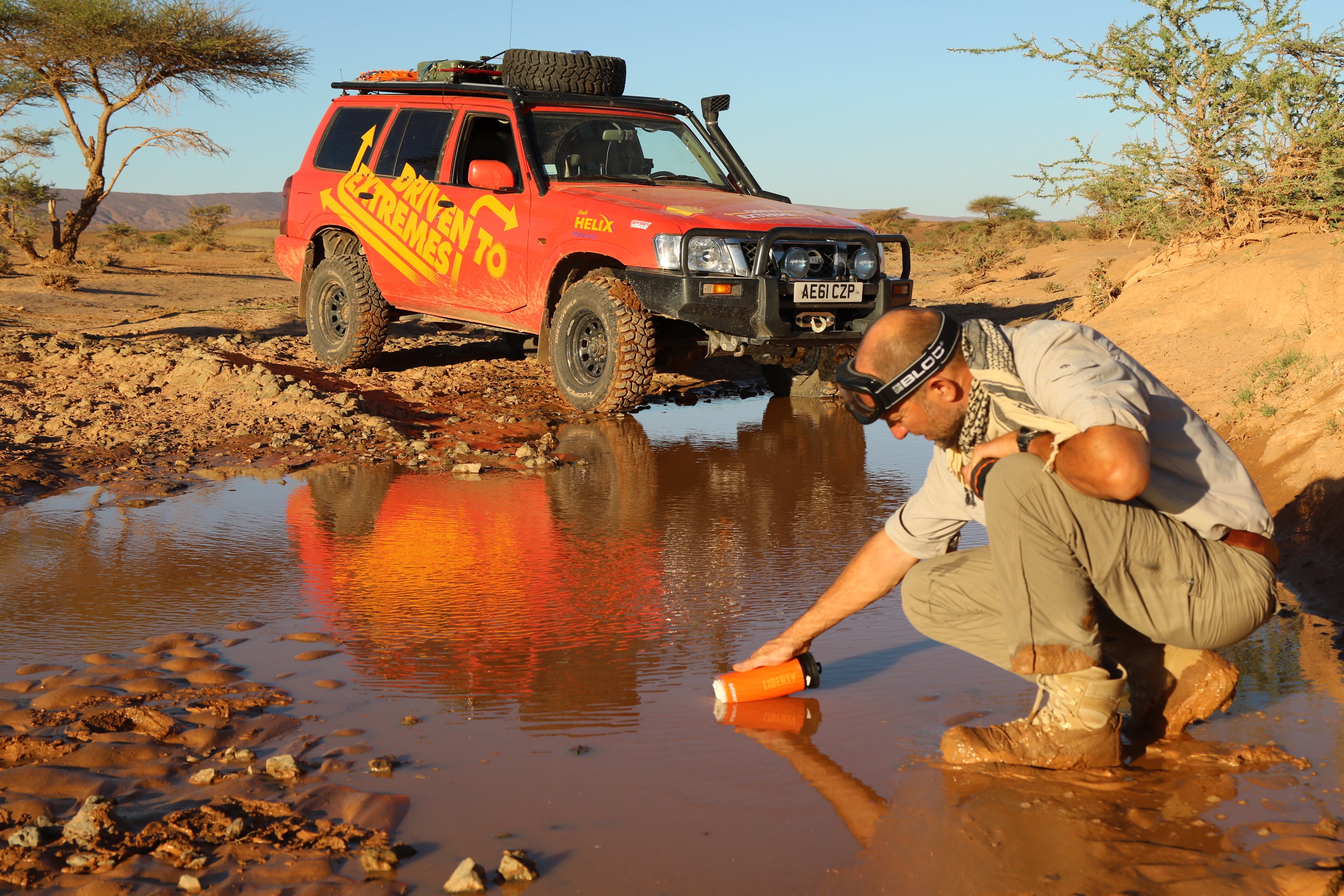 Person refilling LifeSaver Liberty bottle from a muddle puddle by a red off-road vehicle in a desert setting