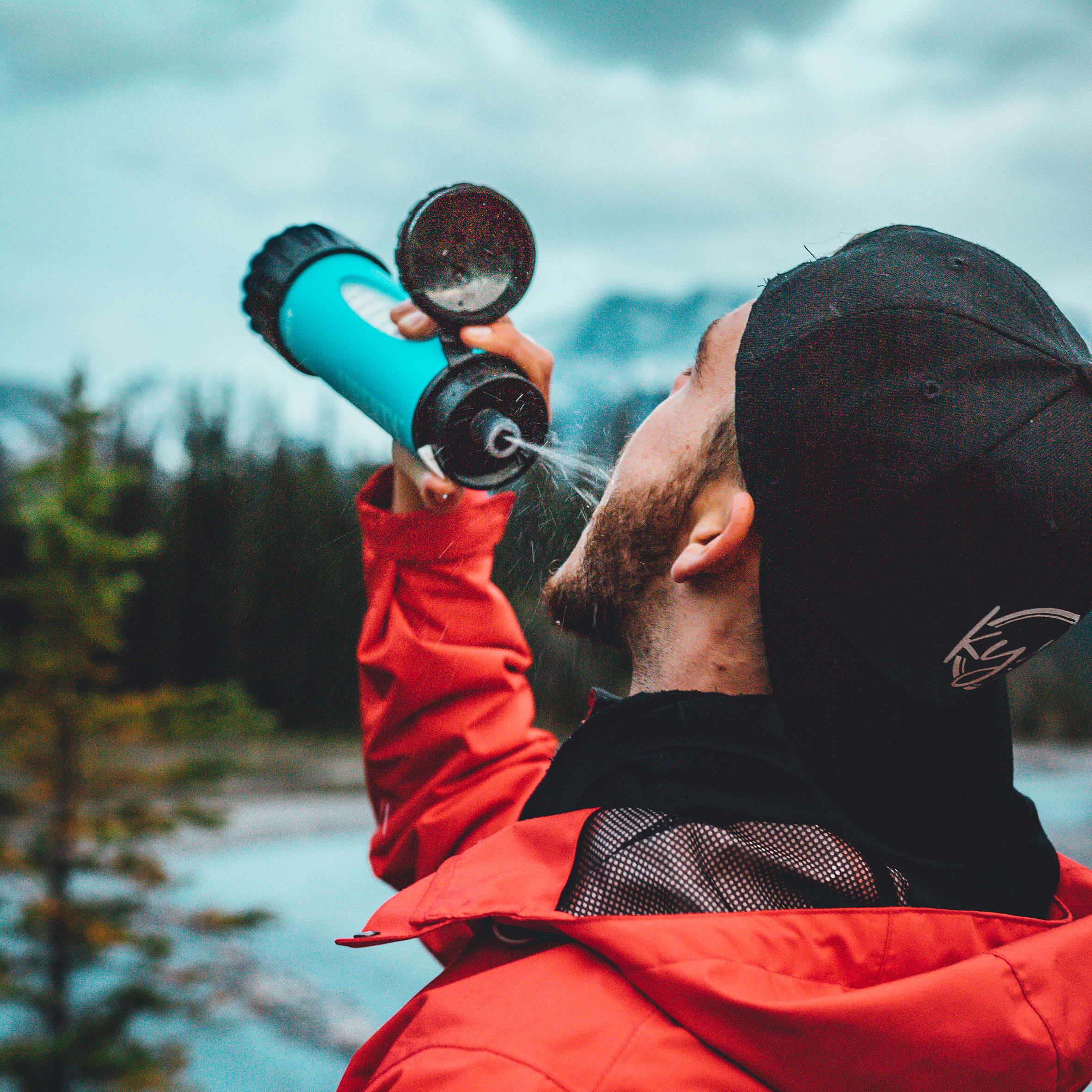 Person in a red jacket drinking from a blue LifeSaver Liberty bottle outdoors with trees and water in the background