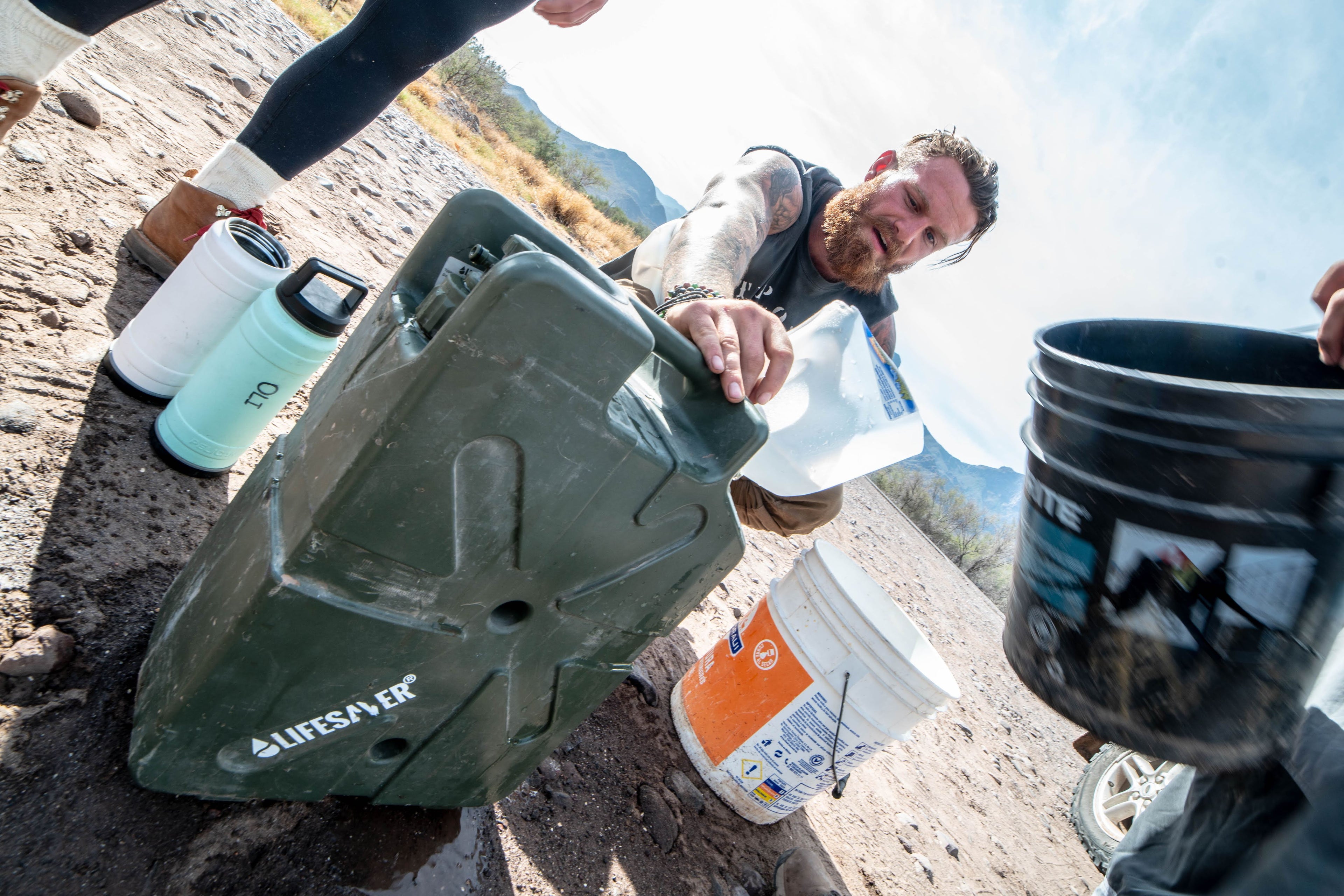 Person using a green jerrycan water filter on a dirt path with mountains in the background