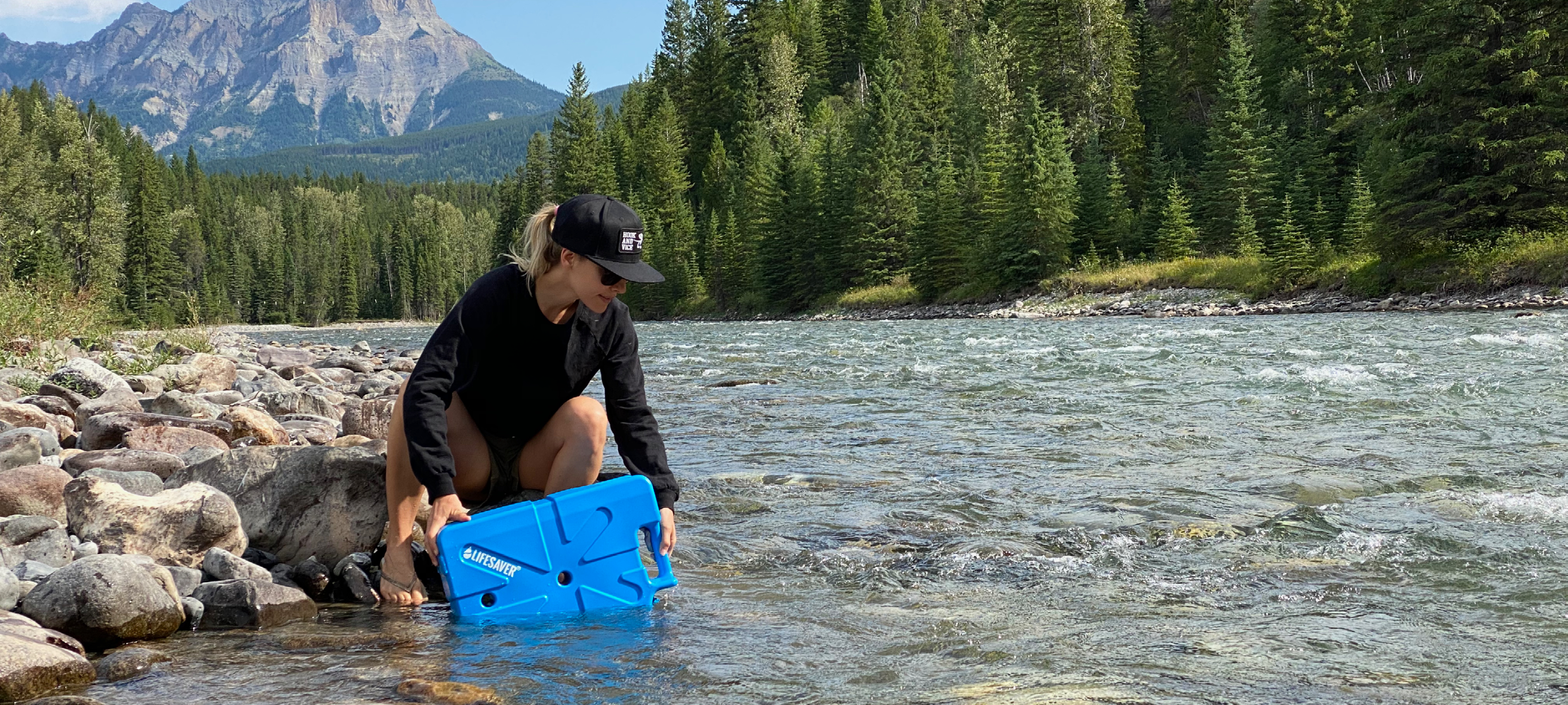Person filling up a blue LifeSaver Jerrycan by a river with mountains in the background