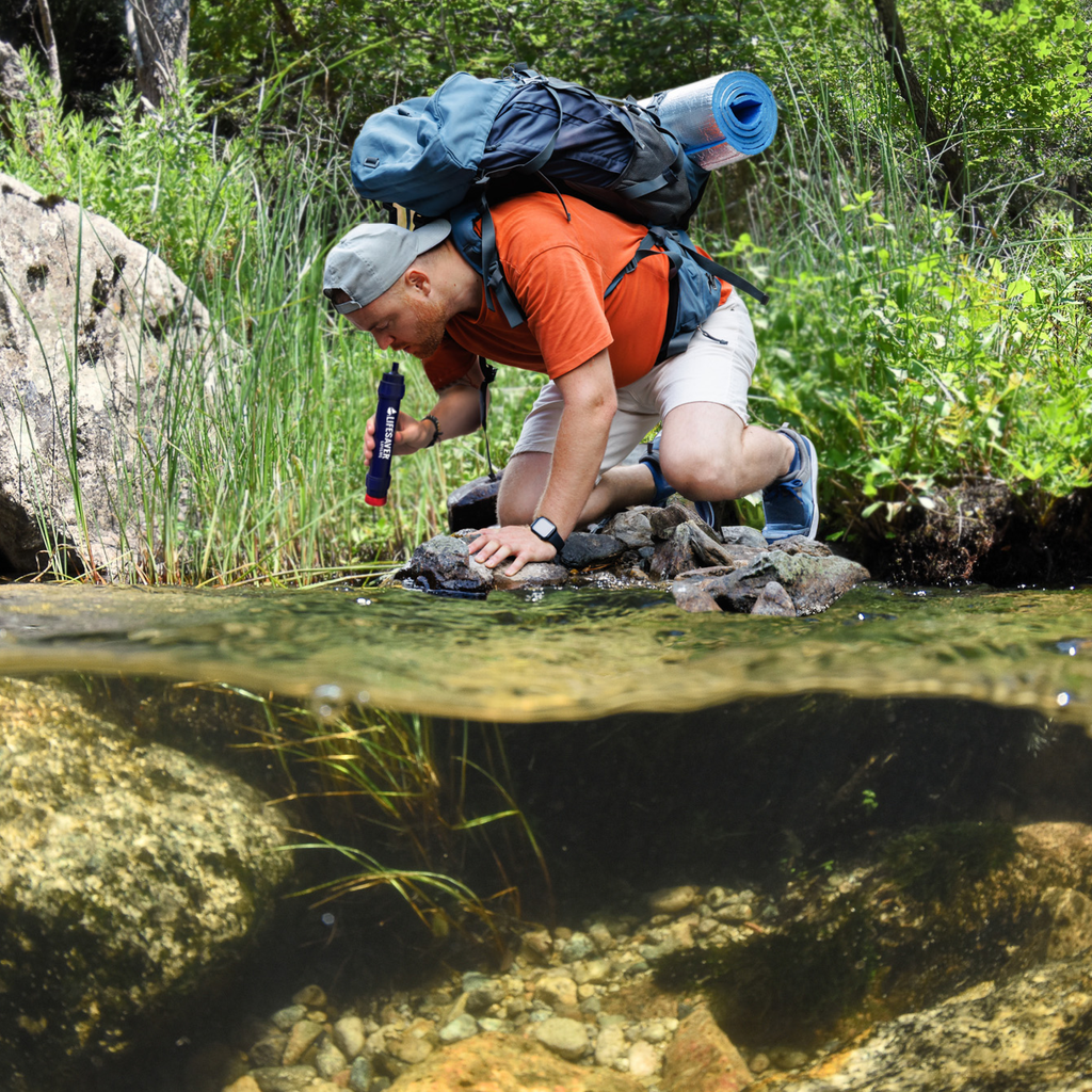 Hiker with backpack crouched beside a stream to drink using a LifeSaver Lifeline filter straw