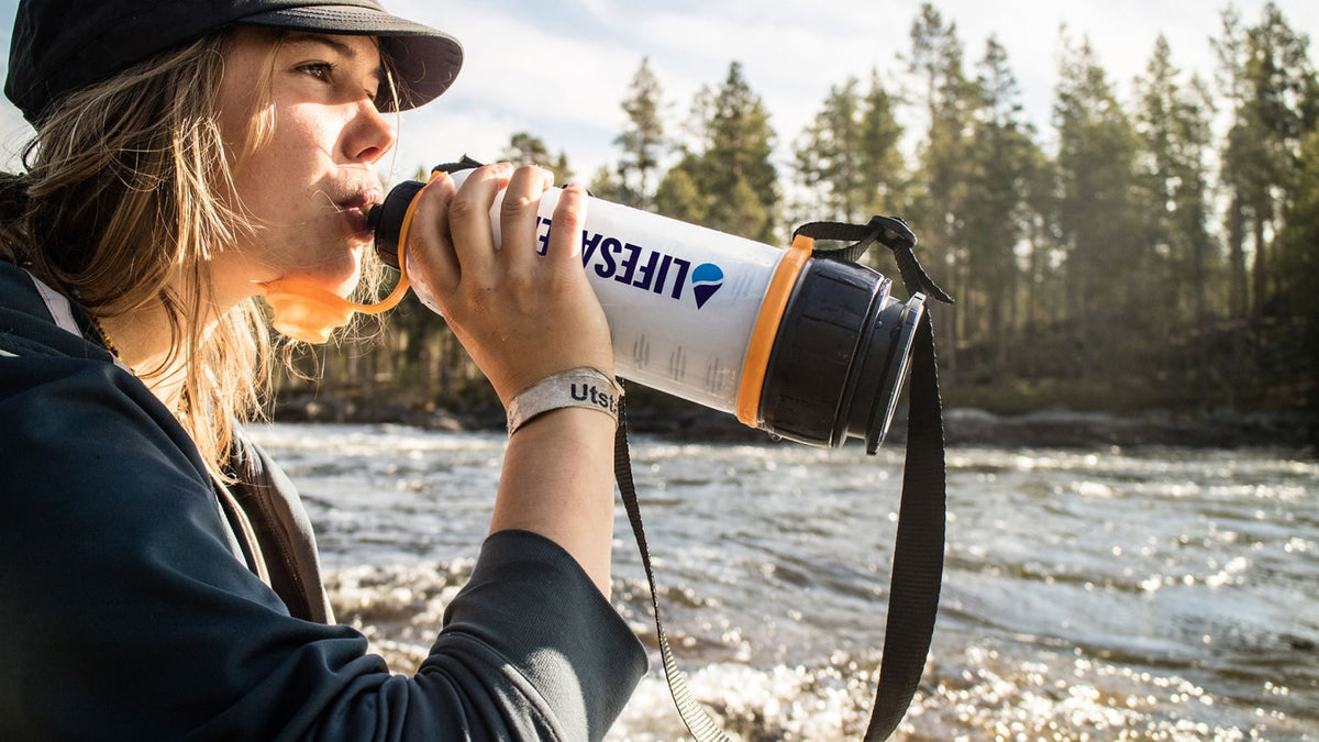 Person drinking from a LifeSaver water filter bottle by a river with trees in the background