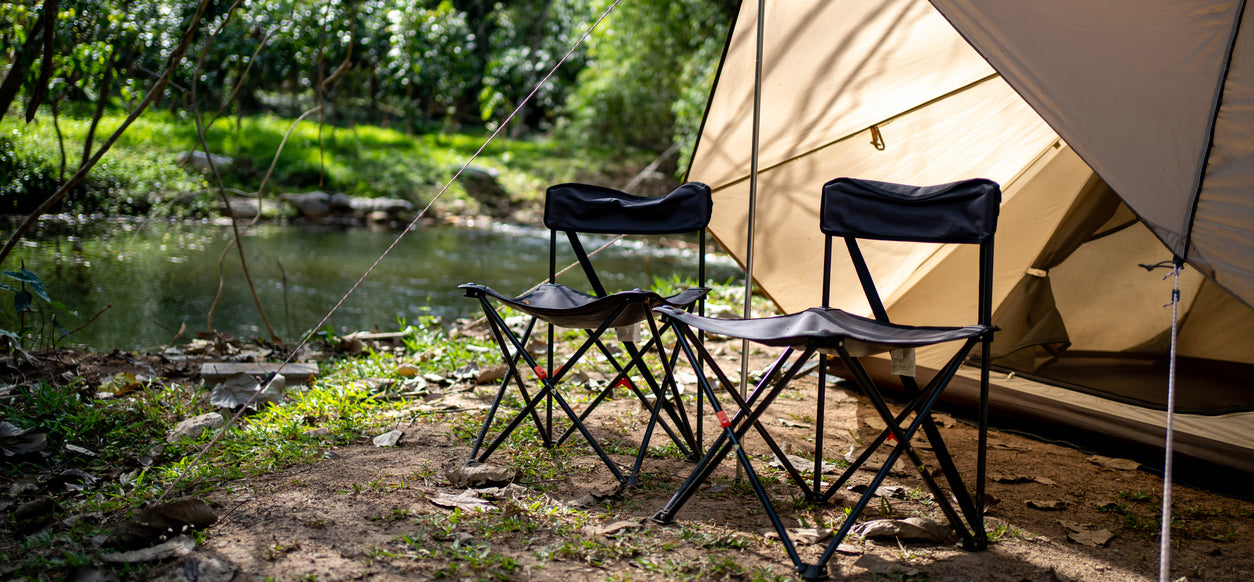 Two black folding chairs in front of a beige tent by a body of water with greenery.